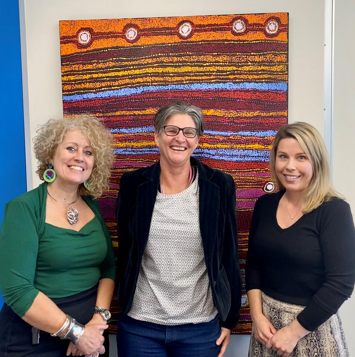 Karen Walsh CEO Venture Housing on the left, Anti-Discrimination Commissioner Sally Sievers and Venture Chief Operations Officer Sarah Thurgood smiling in front of a large colourful Aboriginal artwork.