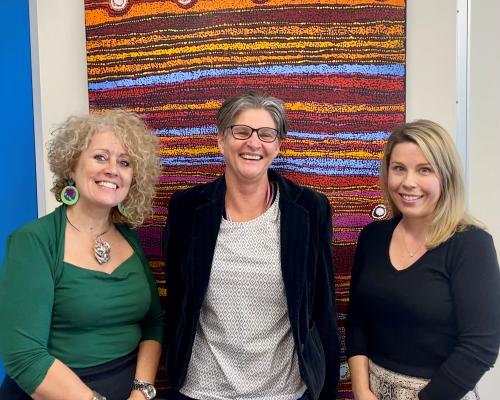 Karen Walsh CEO Venture Housing on the left, Anti-Discrimination Commissioner Sally Sievers and Venture Chief Operations Officer Sarah Thurgood smiling in front of a large colourful Aboriginal artwork. 