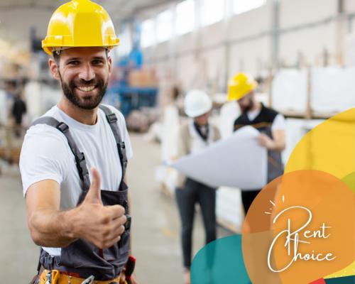 Male construction worker with beard wearing overalls and a yellow hard hat in a warehouse smiling at the camera and giving the thumbs up.